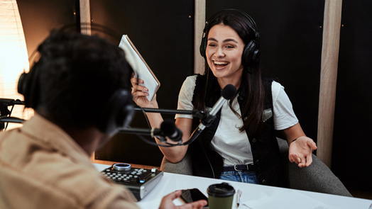 Portait of happy female radio host laughing, listening to male guest, presenter and holding a script paper while moderating a live show in studio