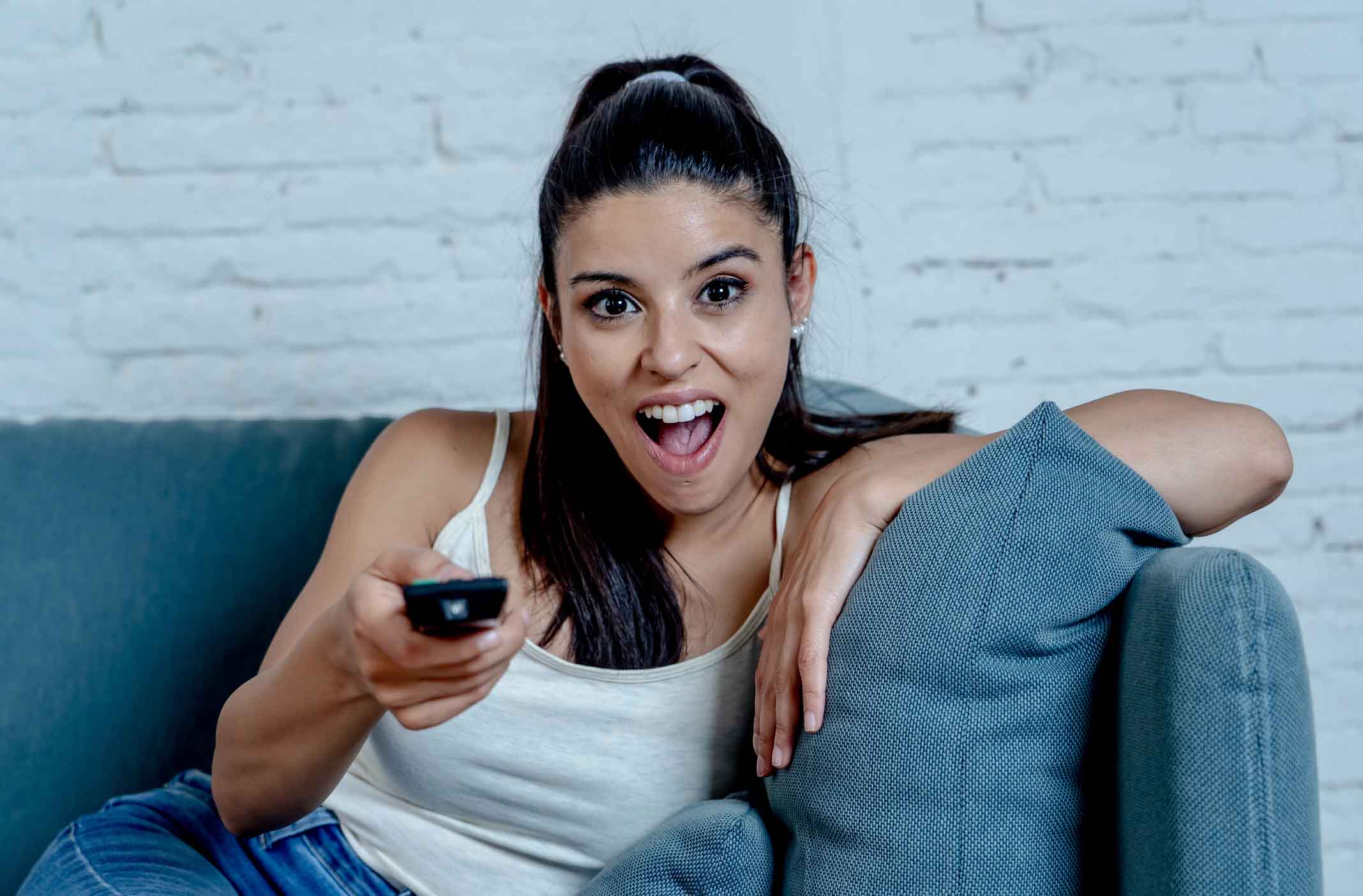 Happy beautiful young latin woman having fun watching TV with control remote in hand sitting on couch at home in television relax lifestyle concept.